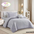 A bedding set displayed on a bed in a styled bedroom scene, with a product inset showing the packaging/label for a 5-piece comforter set. The bedding is a pale lavender-gray color, and the room has beige walls, curtains, and a light wood floor.